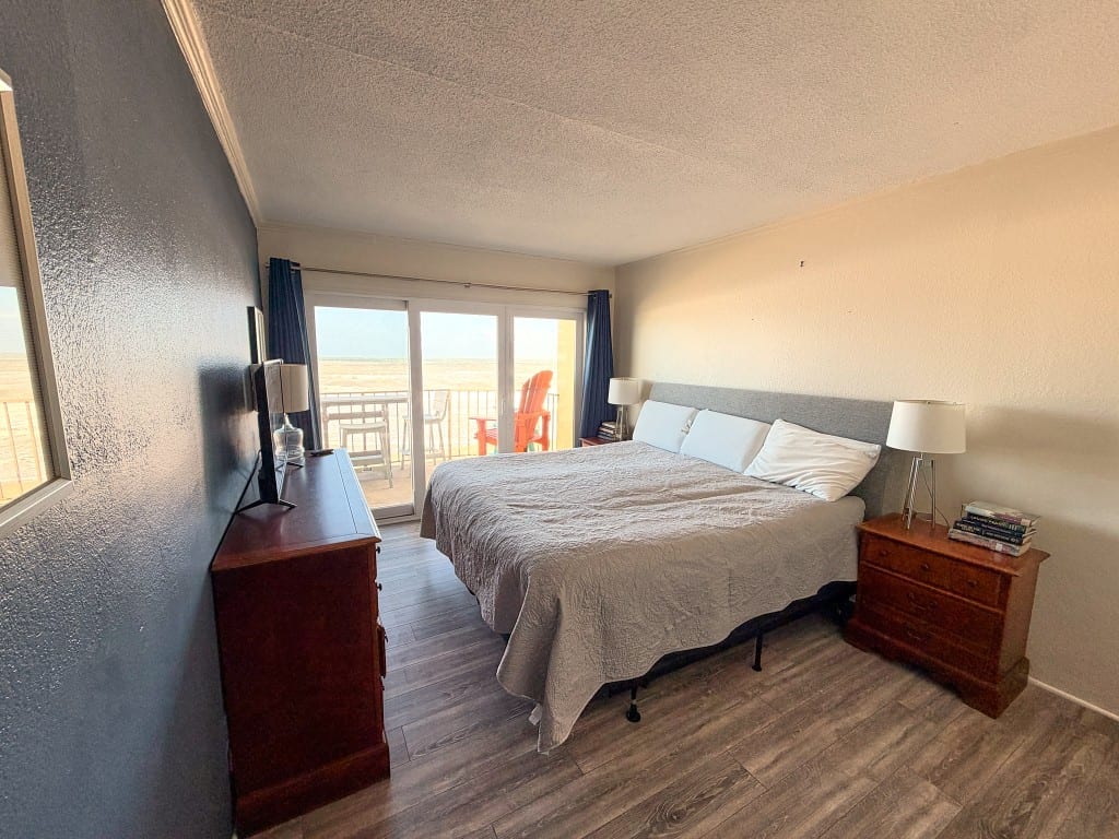King bed with a gray quilt, a wood dresser with a television, and sliding doors opening to the balcony and ocean