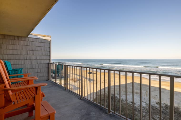 Corner of the top-floor balcony with wooden Adirondack chairs facing the railing, the beach, and open ocean.