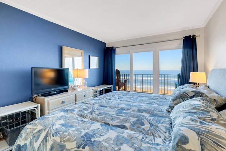 King bed with coastal bedding beside sliding glass doors, with the ocean filling the doorway.
