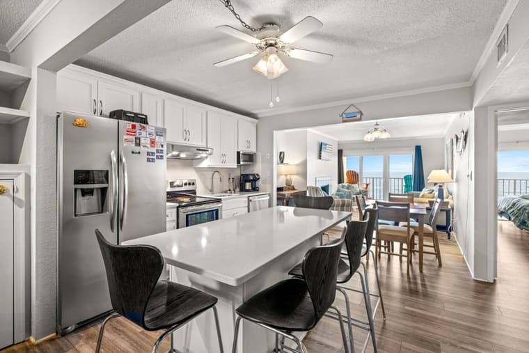 Kitchen with a white island and black stools, stainless steel refrigerator and range, open to the dining area and balcony doors.