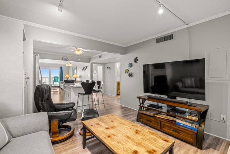 Living room from the entry side showing the television, a leather chair, and the kitchen and balcony doors beyond.